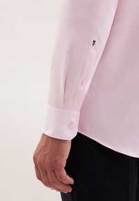 Mano di un uomo accanto a un polsino di camicia a maniche lunghe rosa chiaro con bottoni e un piccolo ricamo di fiore nero, indossando pantaloni neri.
