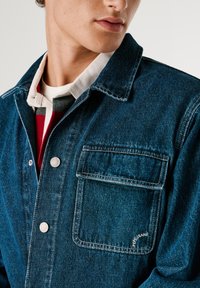 Giubbotto in denim blu scuro, con colletto a punta, chiusura con bottoni e taschino sul petto con cuciture bianche e dettaglio del logo.