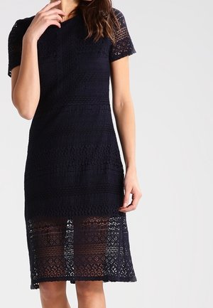 Femme portant une robe noire en dentelle longueur genou avec des manches courtes, présentant une superposition en dentelle transparente de mi-cuisse à l'ourlet.