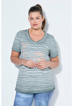 Kvinde med brunt hår i en hestehale bærer en grå stribet V-hals T-shirt med en hval og teksten "Meer geht", kombineret med blå jeans.