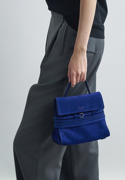 Άτομο που κρατάει μια μπλε σουέντ τσάντα Moschino με λαβή και λεπτομέρεια ζώνης, φορώντας γκρι παντελόνι και μαύρη μπλούζα, σε φωτεινό φόντο.