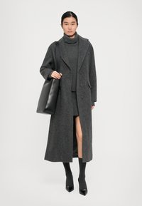 Femme portant un long manteau gris foncé, un pull à col roulé assorti et une jupe, un sac fourre-tout noir, des chaussettes longues et des escarpins noirs à bouts pointus.