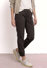 Personne portant un pantalon de jogging noir à cordon, une veste bomber beige, une chemise blanche et des baskets blanc cassé, debout sur un sol clair.