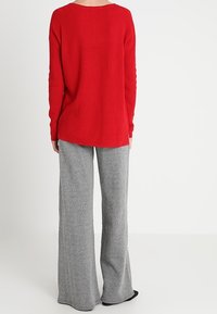 Pull en maille côtelée rouge avec un col large et un dos long, associé à un pantalon évasé en chevron gris. Texture douce ; pas de quincaillerie visible.