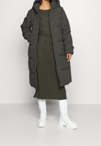 Lång, vadderad olivgrön kappa med huva och fickor fram, bärs över en matchande klänning. Kombinerad med vita snörda kängor.