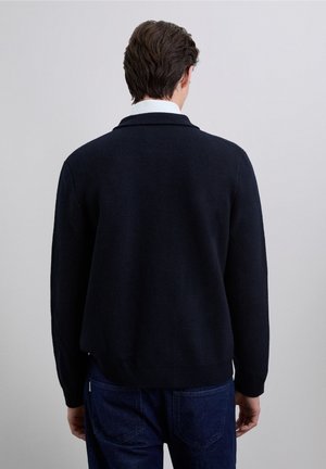 Jersey de punto azul marino con un corte holgado, que cuenta con un pequeño cuello y puños acanalados. La textura es suave, con un sutil patrón tejido.