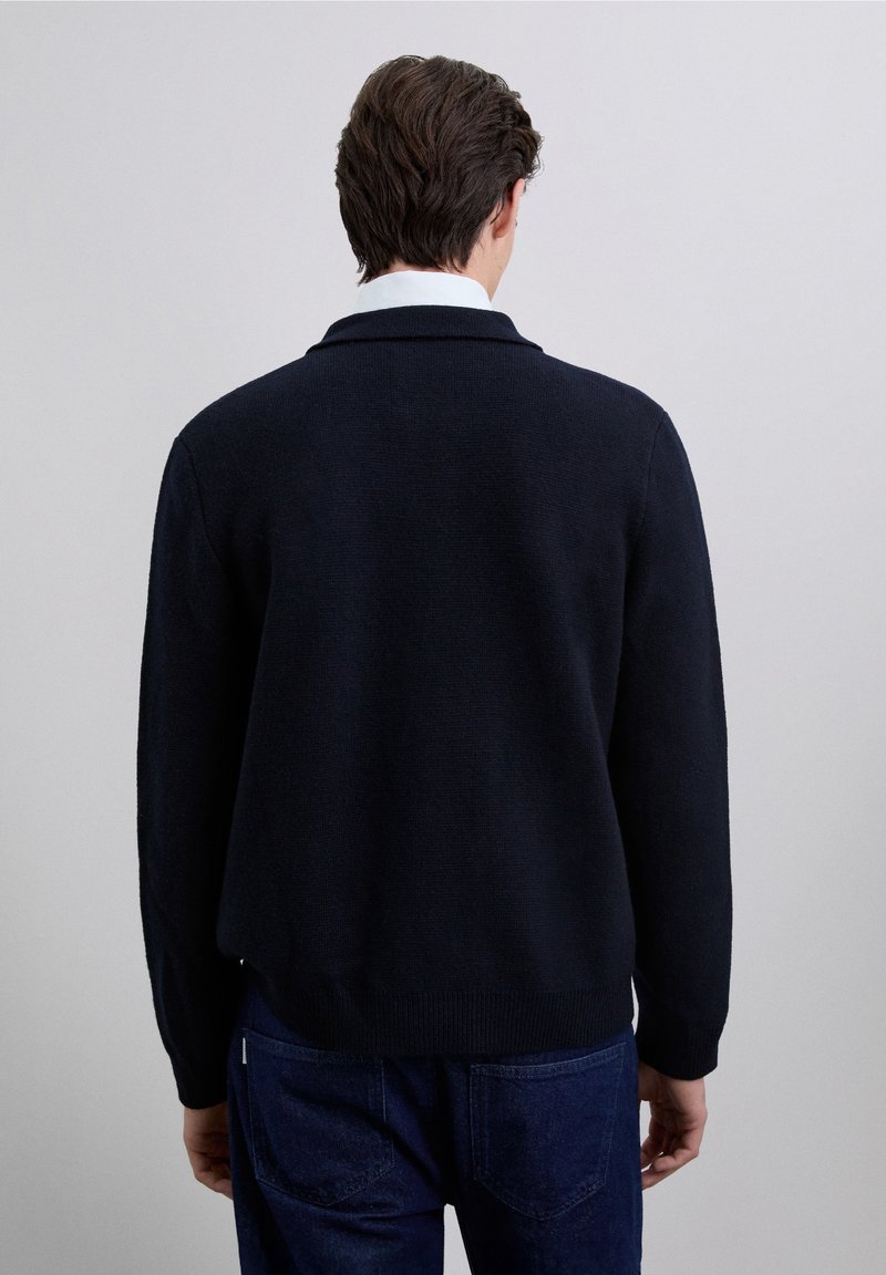 Marinegebroken gebreide trui met een losse pasvorm, voorzien van een kleine kraag en geribde manchetten. De textuur is soepel, met een subtiel geweven patroon.