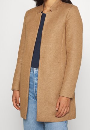 Woman wearing a camel-colored, collarless long coat over a navy top and light blue jeans, standing against a plain white background.