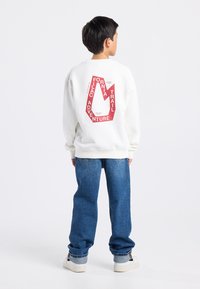 Valkoinen collegepaita, jossa on punainen graafinen printti takana, sisältäen tekstiä ja vuoristosymbolin; yhdistettynä sinisiin löysiä farkkuihin ja valkoisiin tennareihin.