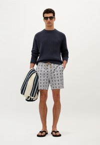 Navy knitted sweater, patterned swim shorts in white and navy, holding a striped towel, black flip-flops, neutral background.