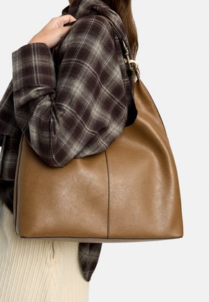 Kobieta ubrana w brązową kratowaną kurtkę i kremowe spodnie, niosąca dużą beżową skórzaną torbę na ramię ze złotymi okuciami.