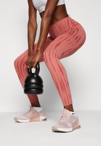 Leggings ajustadas de cintura alta em rosa com padrão vertical, combinados com sapatilhas Nike rosa clara. Um kettlebell preto está sendo levantado.
