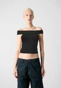 Melna off-the-shoulder blūze, izgatavota no stiepjama auduma ar pieguļošu piegriezumu, īsu garumu un gludu tekstūru. Apvienota ar tumši ziliem kravas biksēm.