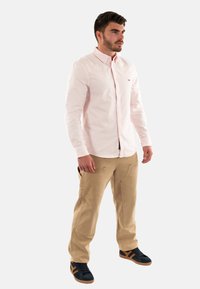 Chemise rayée rose clair avec des boutons, pantalon beige avec poches et baskets noires avec des accents marron. Le modèle se tient de profil à gauche.