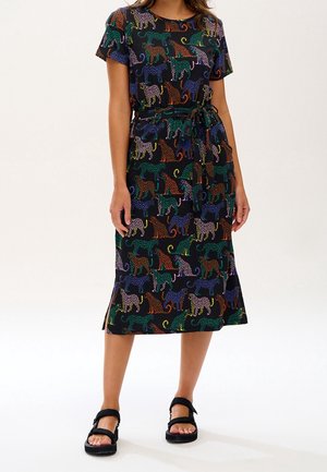 Kobieta w czarnej sukience midi z kolorowym nadrukiem w cętki, krótkimi rękawami, wiązaniem w talii i czarnych sandałach z otwartymi palcami.