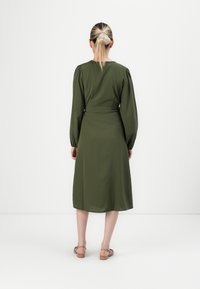 Woman with blonde hair in a ponytail wearing an olive green, long-sleeve, midi dress and brown sandals, standing against a white background.