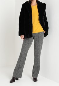 Manteau en fausse fourrure noir sur une chemise à manches longues jaune, associé à un pantalon à motifs gris et des bottes à talons noires brillantes.