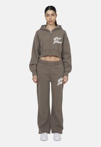 Felpa corta marrone con zip e testo bianco, abbinata a pantaloni a vita alta coordinati. Il tessuto sembra morbido con un taglio rilassato. Sono indossate sneakers bianche.