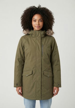 Vrouw met krullend haar die een olijfgroene parka-jas draagt met bontkraag aan de capuchon, een ritszak op de borst en klepzakken, staand tegen een effen achtergrond.