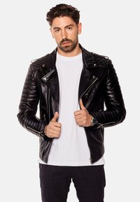 Svart läderbikerjacka med kviltade axlar, silverdragkedjor och en figurnära design. Buren över en vit t-shirt, visad med tummen upp.
