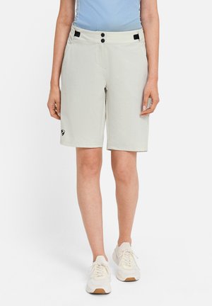 Person trägt hellgraue kniehohe Shorts mit schwarzen Knopfdetails und weißen Turnschuhen, steht vor einem schlichten Hintergrund.