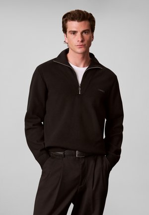 Jeune homme portant un pull noir à demi-fermeture éclair par-dessus une chemise blanche, pantalon noir et ceinture noire, debout, les mains dans les poches, sur fond gris.