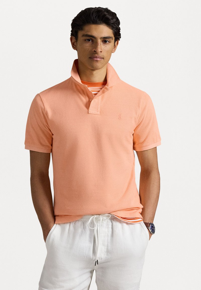 Polo a maniche corte in cotone di un arancione chiaro con colletto e dettaglio del logo. Indossato sopra una camicia a righe, abbinato a pantaloni bianchi.