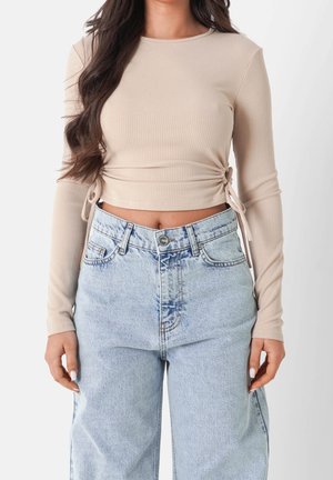 Frau trägt ein beiges, langärmliges, kurz geschnittenes, geripptes Oberteil mit seitlichen Bändern und hellblauen, hoch taillierten Jeans, steht vor einem weißen Hintergrund.