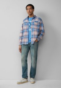 Karierter Hemd in Blau, Pink und Weiß; blaues Grafik-T-Shirt darunter; helle Denim-Jeans; weiße Sneaker mit beigen Akzenten.