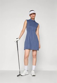 Niebieska golfowa sukienka bez rękawów z delikatnym falistym wzorem, z kołnierzykiem i rozkloszowaną spódnicą; w połączeniu z białymi sportowymi butami i rękawiczkami.