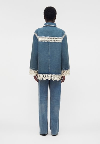 Άτομο φορά μπλε τζιν μπουφάν και παντελόνια με λευκές λεπτομέρειες από φλοράλ δαντέλα στα μανίκια του μπουφάν, στην πλάτη και στο τελείωμα, πάνω σε απλό φόντο.