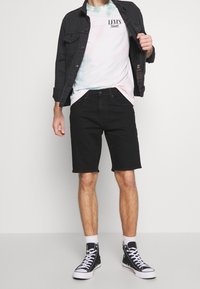 Pantaloni corti in denim nero e una maglietta tie-dye, sopra una giacca in denim nero. Sneakers alte nere completano l'outfit.