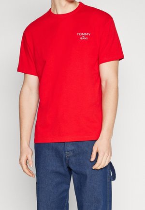 Mann trägt rotes Tommy Jeans T-Shirt und blaue Jeanshose, steht vor schlichtem hellem Hintergrund.