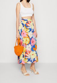 Maxihame kukumaa bluu yenye maua makubwa ya pinki, njano, na rangi ya machungwa; imeunganishwa na koti ya juu nyeupe, viatu vya beige, na begi la kushona la rangi ya machungwa.