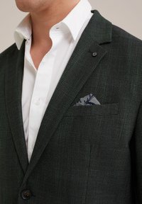 Zaļš raibs bleizeris ar teksturētu audumu, ar vienas pogas aizdari un rakstainu kabatas lakatiņu, kas ievietots krūškurvja kabatā.