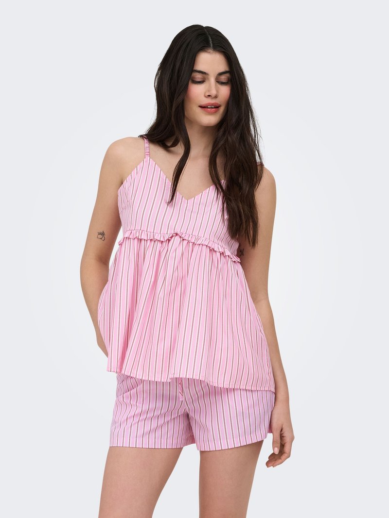 Femme aux longs cheveux sombres portant un haut sans manches rayé rose et blanc avec volant et un short assorti, debout avec une main dans la poche.