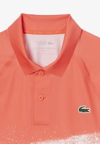 Korallfärgad polo med slim fit, som har en klassisk krage och tre knappar. Lacoste-logotyp i grönt och rött på vänster sida.