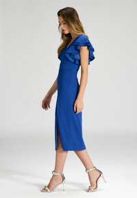 Robe bleue avec des manches à volants, silhouette ajustée et fente haute sur la cuisse, associée à des sandales à talons hauts argentés. Texture de tissu lisse.