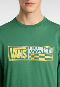 Grünes Langarm-T-Shirt aus Baumwolle mit einem auffälligen gelb-weißen "VANS OFF THE WALL" Grafikdesign und einem karierten Muster.