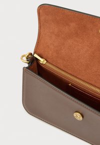 Sac en cuir marron avec rabat ouvert montrant l'intérieur en daim, une poche zippée en or et une fermeture à bouton-pression ronde en or gravée du nom de la marque.