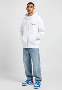 Felpa bianca con zip e cordini, caratterizzata da un logo in scrittura nera. Indossata con jeans azzurri e sneaker blu e bianche.