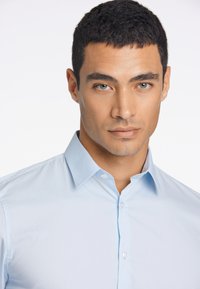 Camicia button-up azzurra chiara realizzata in tessuto liscio, con colletto a punta e patta dei bottoni standard. Vestibilità su misura con minimal texture.