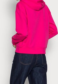 Felpa rosa con cappuccio, maniche lunghe e orlo a coste, abbinata a jeans in denim blu scuro con tasche e cuciture arancioni.