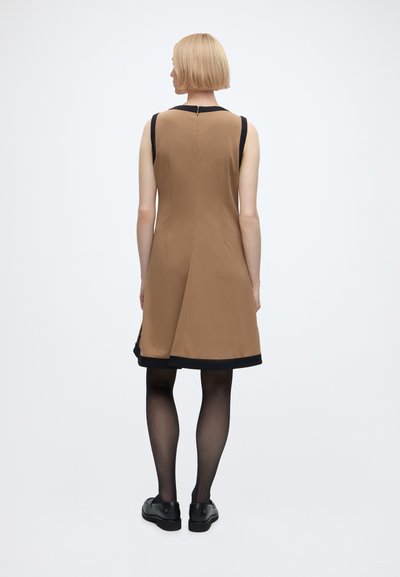 Femme aux cheveux blonds courts portant une robe marron sans manches avec bordure noire, des collants noirs et des chaussures noires, debout de dos.