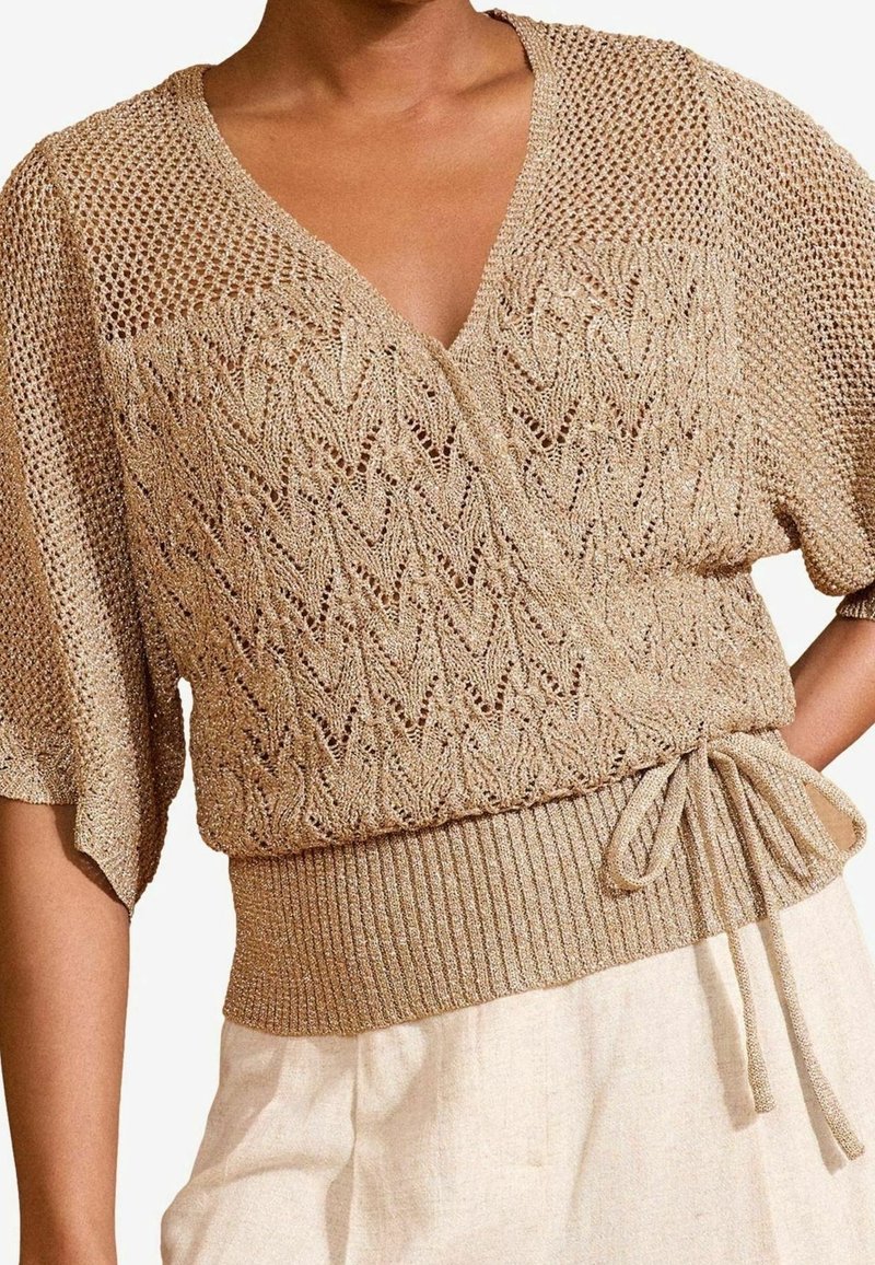 Beżowy sweter w formie nawijania z dekoltem w serek, krótkimi rękawami, otwartym wzorem splotu i prążkowanym dołem. Detal w postaci wiązania w talii nadaje mu struktury.