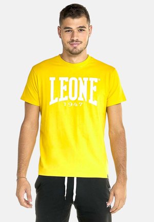 Mann mit kurzen dunklen Haaren, der ein gelbes T-Shirt mit weißem Text ""LEONE 1947"" und schwarze Hosen mit weißen Kordeln trägt.