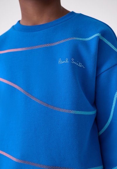 Person wearing a blue Paul Smith sweatshirt with curved, multicolored reflective stripes across the chest and sleeve.