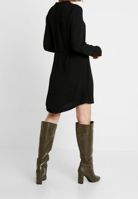 Schwarzes Langarmkleid mit lockerer Passform und abgerundetem Saum, kombiniert mit kniehohen, hochhackigen olivgrünen Stiefeln mit strukturierter Oberfläche.