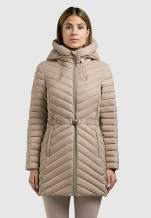Kobieta ubrana w beżowy pikowany płaszcz z kapturem, przednim zamkiem błyskawicznym, paskiem i bocznymi kieszeniami, stojąca na tle gładkiego, jasnego tła.