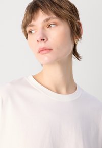 Vit bomullströja med rund halsringning, med en mjuk tygtextur. Subjektet har kort hår och bär en guldring som örhänge.